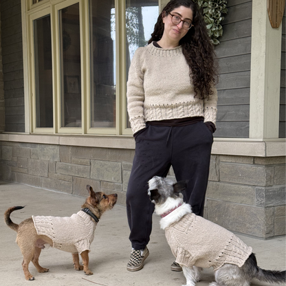 A woman stands on a porch wearing a cream cable-knit sweater, with two small dogs also wearing matching cream knit sweaters. The setting is outdoors in front of a house with gray siding and large windows.