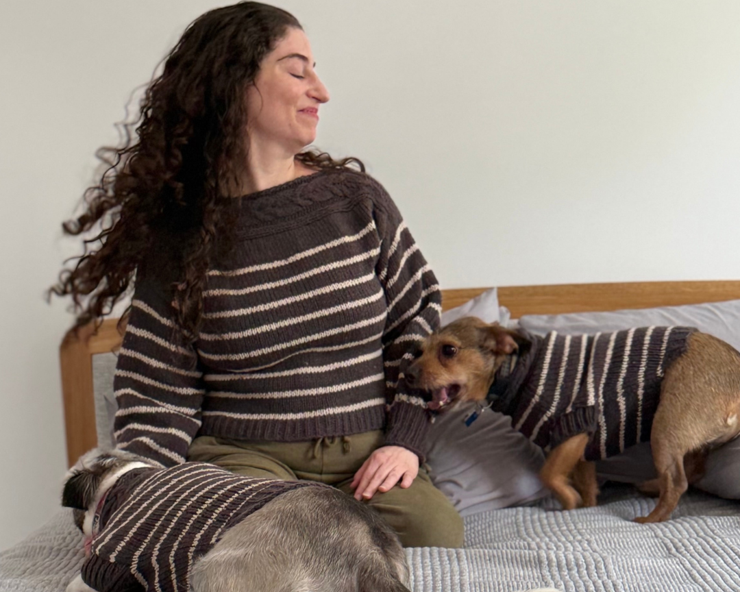 A woman sits on a bed wearing a gray and beige striped knit sweater, with two dogs also wearing matching striped sweaters in a bedroom setting.