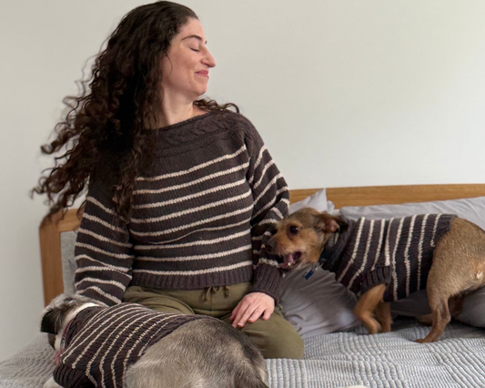 A woman sits on a bed wearing a gray and beige striped knit sweater, with two dogs also wearing matching striped sweaters in a bedroom setting.
