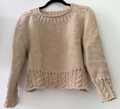 A beige knit cropped sweater with textured cable and rib patterns, hanging on a black hanger against a plain white wall.