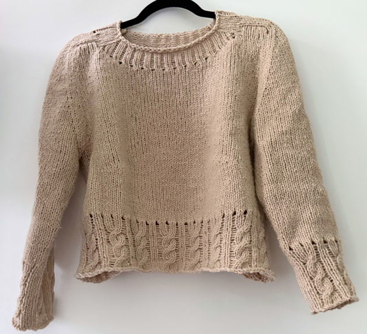 A beige knit cropped sweater with textured cable and rib patterns, hanging on a black hanger against a plain white wall.